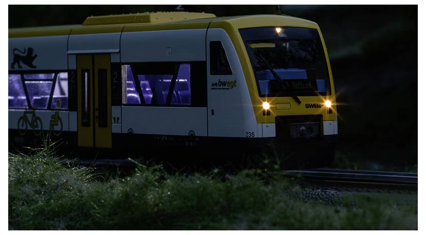 Ein kleines Modell-Elektrozug, gelb und weiß, beleuchtet, fahrend auf Miniaturgleisen durch eine grüne Landschaft.