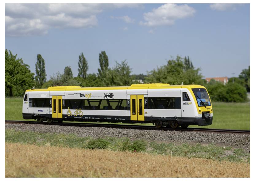 Ein gelb-weißer Zug fährt auf einer Schiene durch ein grünes Gebiet mit Bäumen und Wolken im Hintergrund.