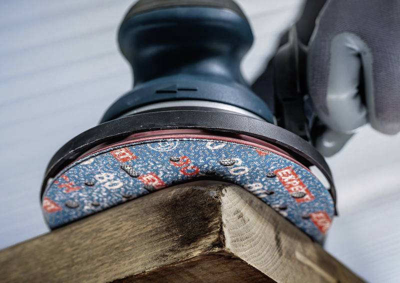 Eine elektrische Schleifmaschine bearbeitet die Ecke eines Holzbretts für eine glatte Oberfläche. Eine behandschuhte Hand hält die Maschine.