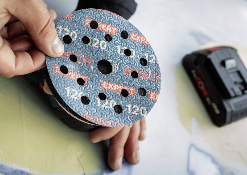 Eine Person hält eine Schleifpapier-Scheibe mit einer blauen und roten Musterung in der Hand, bereit zum Anbringen an ein Elektrowerkzeug.