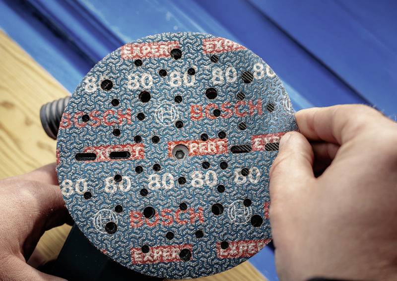 Zwei Hände montieren eine Schleifscheibe mit der Aufschrift 'Bosch 80' auf eine Schleifmaschine. Hintergrund aus Holz und Blau.