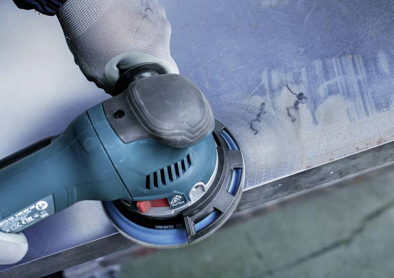 Eine Person schleift mit einer blauen, elektrischen Schleifmaschine eine Metalloberfläche. Handschuh ist sichtbar.