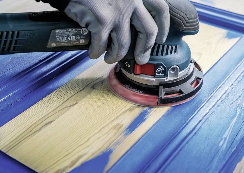 Eine Person schleift mit einem elektrischen Schwingschleifer blauen Lack von einer Holzoberfläche ab, um das Holz freizulegen.