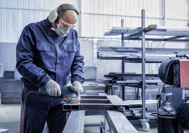 Ein Arbeiter in Schutzkleidung schleift Metall in einer Fabrikumgebung. Im Hintergrund sind Regale mit weiteren Metallteilen sichtbar.