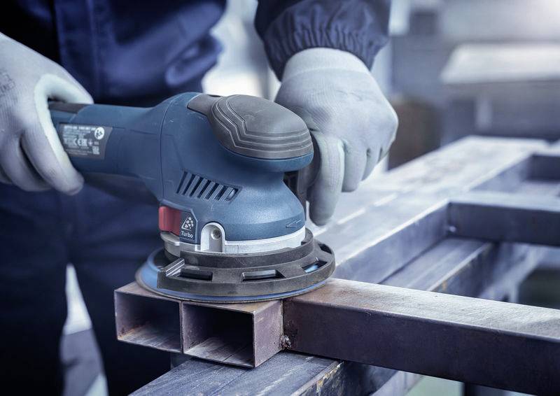 Person schleift Metall mit einer blauen elektrischen Schleifmaschine auf einer Baustelle. Trägt Handschuhe zum Schutz.