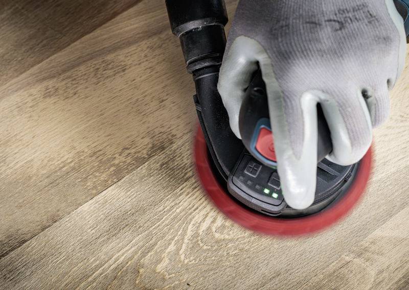 Eine Person schleift mit einem Elektroschleifer eine Holzoberfläche. Der Handschuh schützt die Hand vor Vibrationen und Staub.