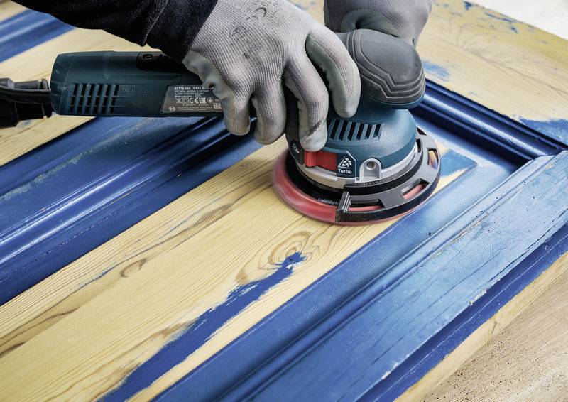 Eine Person schleift eine Holztür mit einer elektrischen Schleifmaschine ab. Die Tür hat blaue Farbreste, die entfernt werden.