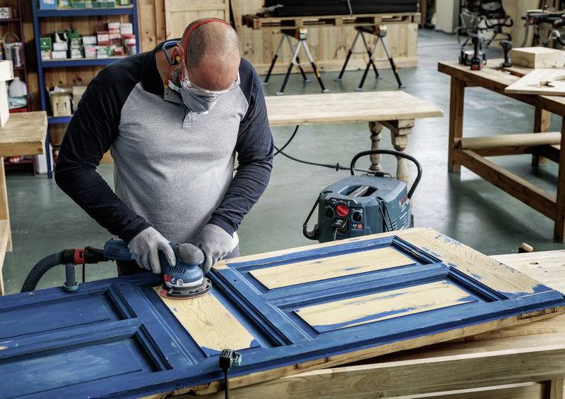 Ein Mann mit Schutzbrille schleift eine blaue Tür in einer Werkstatt. Im Hintergrund sind Werkzeuge und Werkbänke zu sehen.
