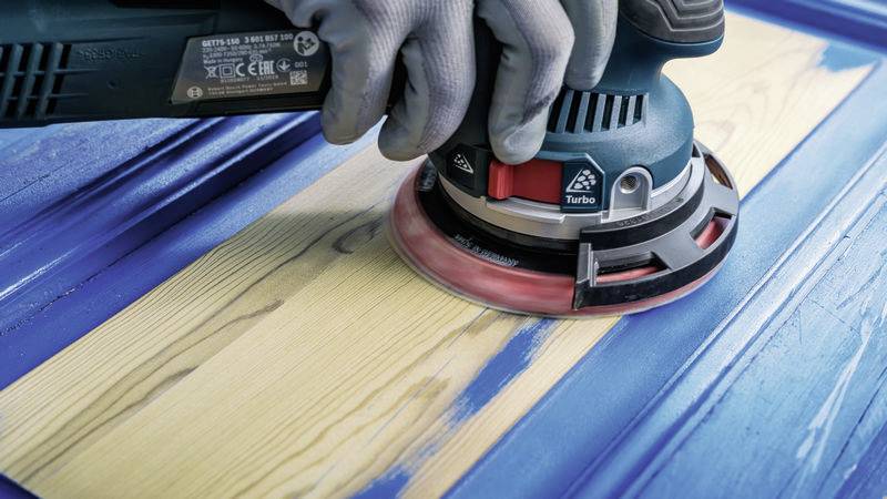 Eine Person schleift mit einem blauen Elektroschleifer eine lackierte Holzoberfläche, um den blauen Anstrich teilweise zu entfernen.