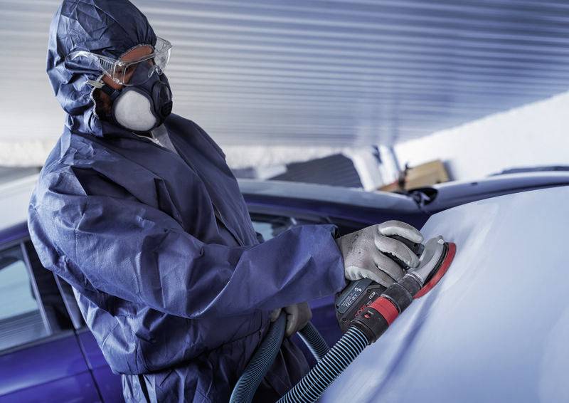 Person in Schutzanzug schleift Autooberfläche. Staubmaske und Schutzbrille werden getragen. Auto in Werkstattumgebung.
