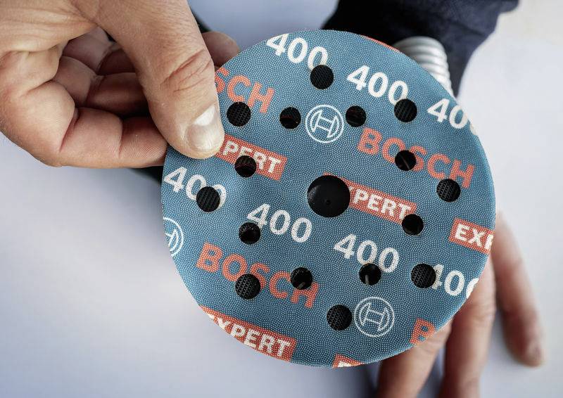 Hand hält eine runde Schleifscheibe mit dem Aufdruck 'Bosch Expert 400'. Die Scheibe hat mehrere Löcher und ist blau gefärbt.