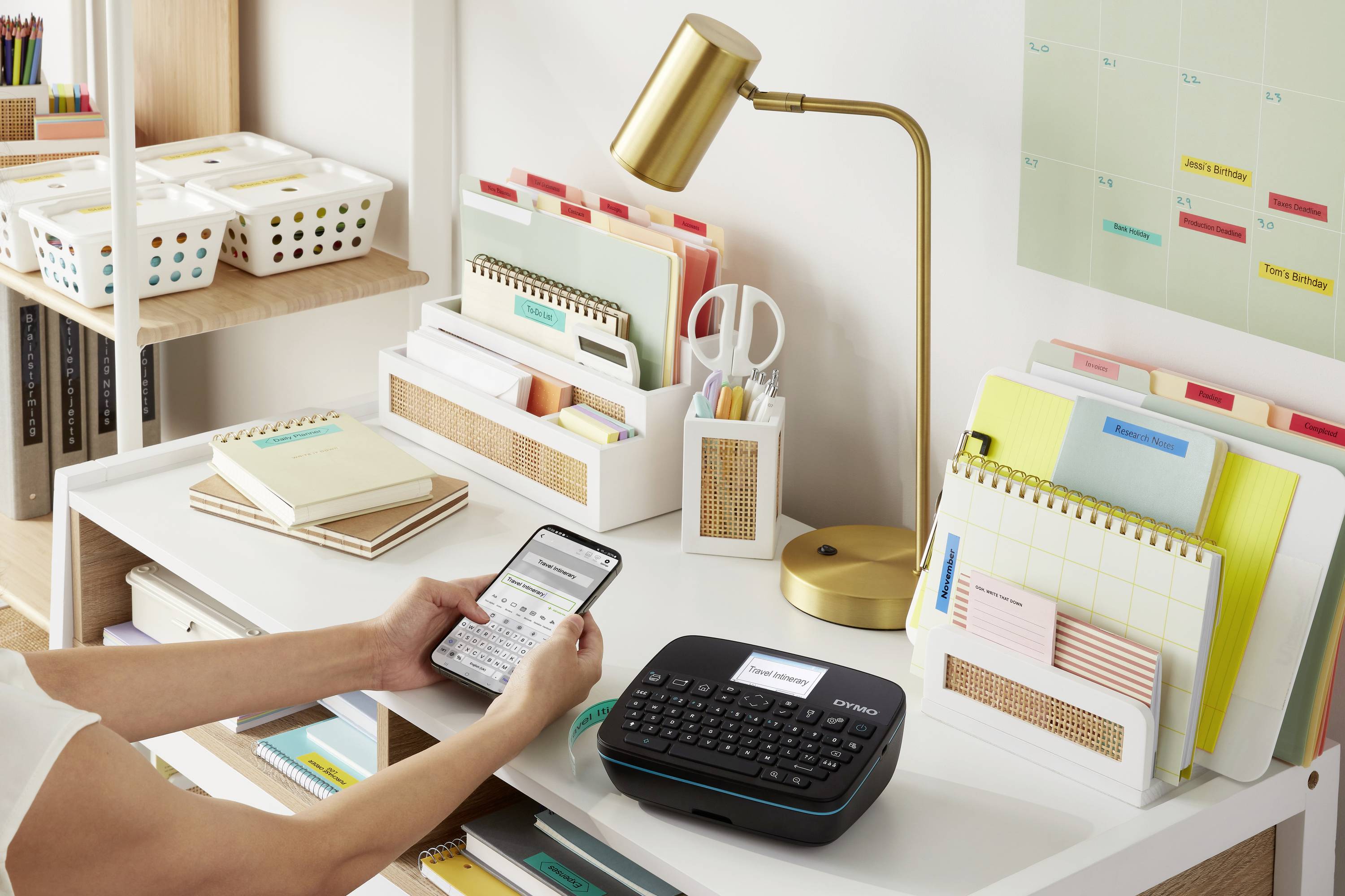 Eine Person bedient ein Etikettiergerät auf einem Schreibtisch, umgeben von organisiertem Büromaterial, einer Lampe und einem Kalender.