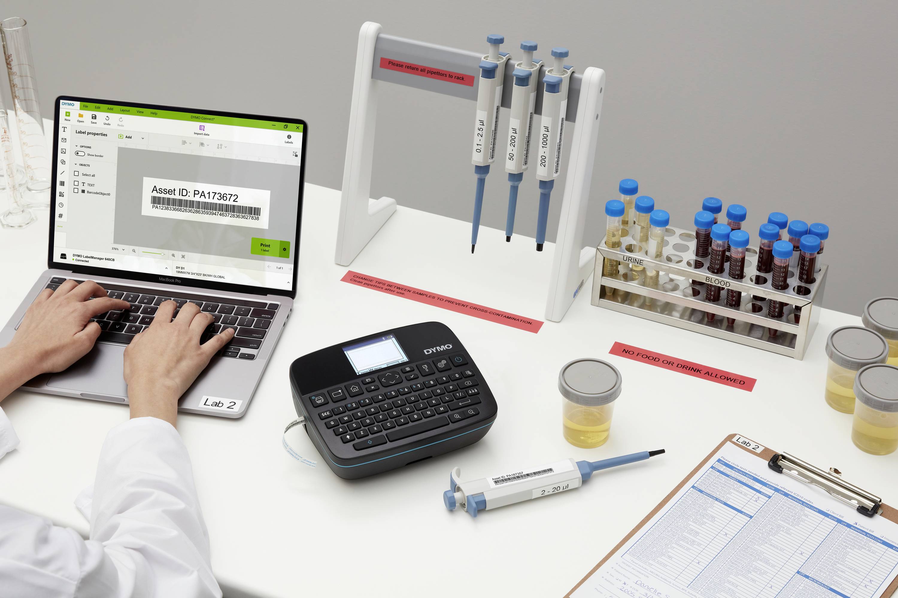 Ein Arbeitsplatz im Labor mit Laptop, Beschriftungsgerät und Probenröhrchen. Eine Person arbeitet am Laptop.