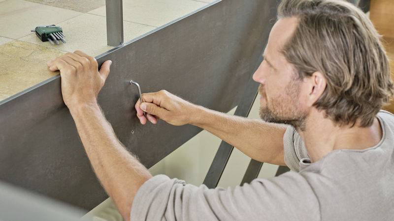 Ein Mann befestigt eine Metallplatte mit einem Imbusschlüssel. Im Hintergrund liegt ein Werkzeug auf dem Boden.