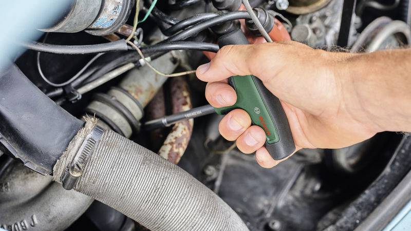 Nahaufnahme einer Hand, die einen Schraubenzieher benutzt, um an einem Autoteil zu arbeiten. Zahlreiche Kabel und Rohre sind sichtbar.