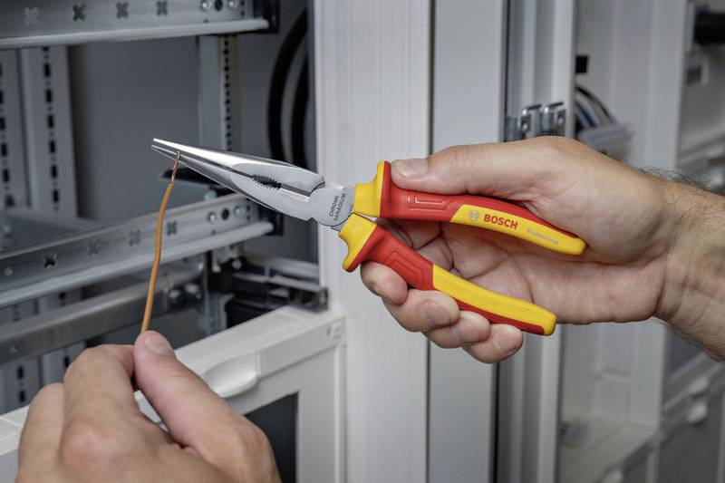 Eine Person hält eine rote-gelbe Kombizange und biegt ein Kabel vor einem offenen Schaltschrank.