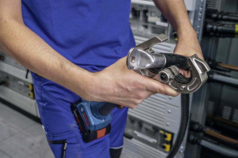 Person in blauer Arbeitskleidung hält ein Werkzeug für Rohrverbindungen in einem technischen Raum. Hintergrund zeigt verschiedene Geräte und Kabel.