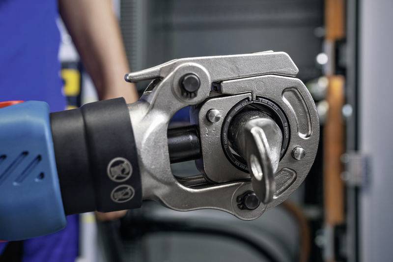 Nahaufnahme eines Werkzeugs, das von einer Person gehalten wird. Es scheint sich um ein elektrisches Gerät zum Klemmen oder Verbinden zu handeln. Im Hintergrund verschwommen ist ein Schrank oder eine Maschine zu sehen.