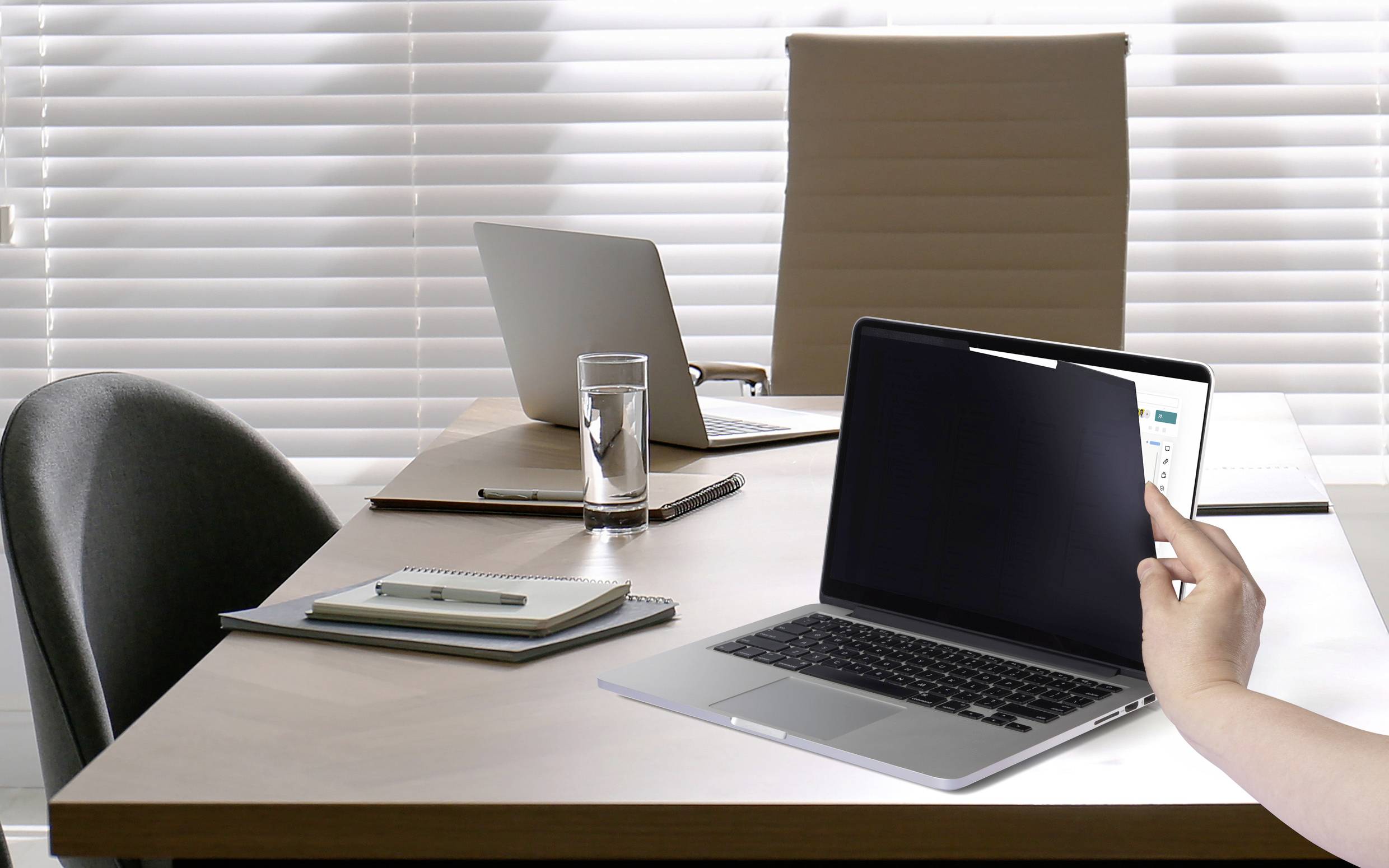 Ein Büro mit drei Laptops auf einem Tisch, daneben ein Glas Wasser und Schreibmaterial. Eine Person hält einen Laptopbildschirm.