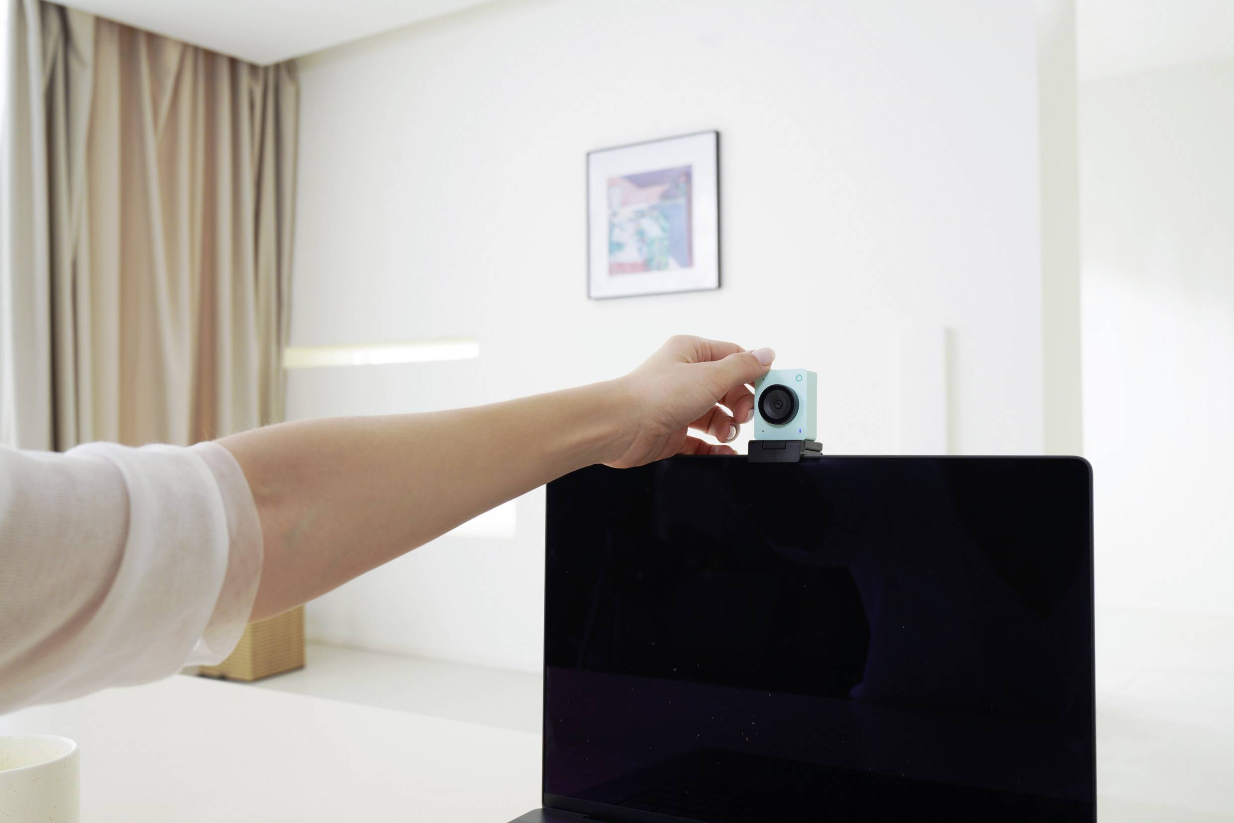 Eine Person platziert eine kleine Kamera auf einem Laptop-Bildschirm in einem hellen Raum mit einem Bild an der Wand im Hintergrund.