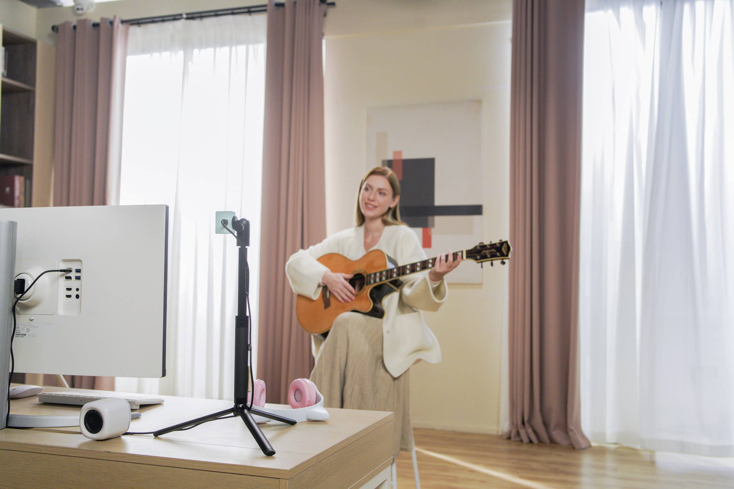 Eine Person sitzt in einem hellen Raum und spielt Gitarre vor einer Kamera auf einem Stativ, was auf ein Online-Streaming hindeutet.