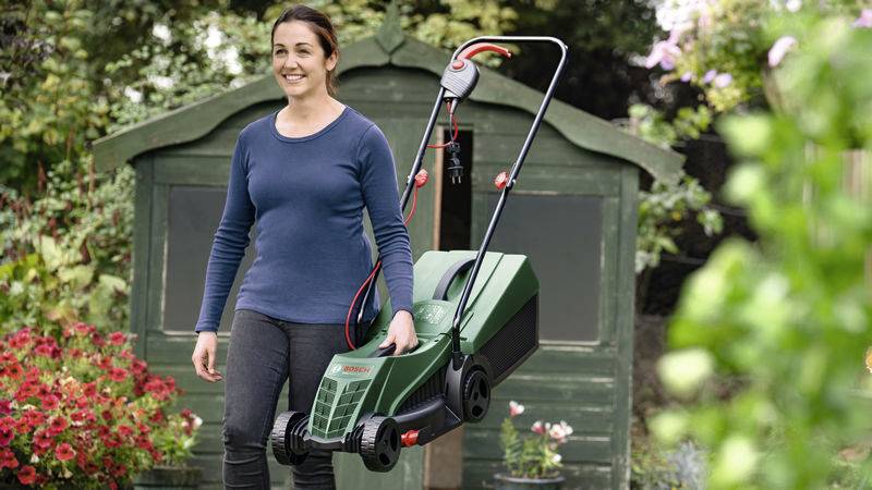 Eine Person steht lächelnd in einem Garten und hält einen grünen Rasenmäher vor einem Schuppen. Im Hintergrund sind Blumen zu sehen.