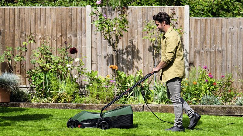 Ein Mann mäht den Rasen im Garten mit einem elektrischen Rasenmäher. Im Hintergrund sind ein Holzzaun und blühende Pflanzen sichtbar.