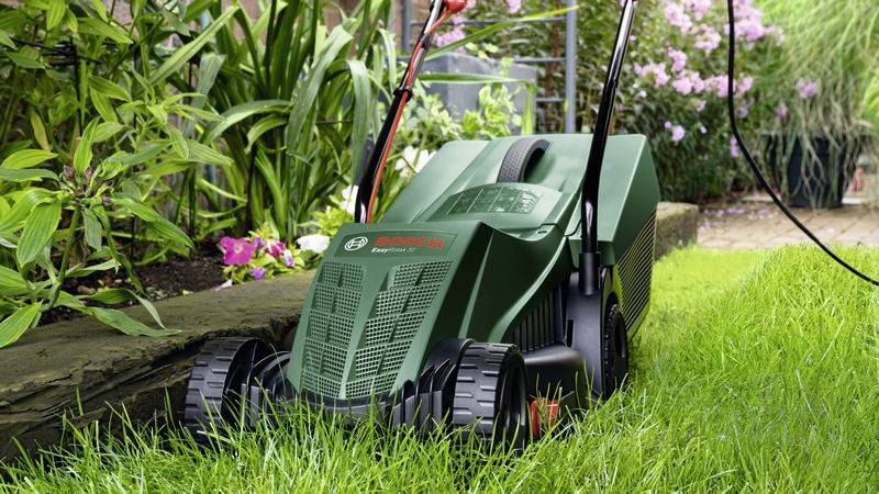 Ein Rasenmäher der Marke Bosch schneidet grünes Gras in einem Garten. Links sind grüne Pflanzen und rechts blühende Sträucher zu sehen.
