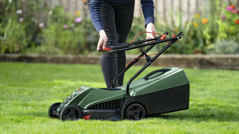 Eine Person mäht Rasen mit einem dunkelgrünen Rasenmäher in einem Garten. Im Hintergrund sind Blumenbeete zu sehen.