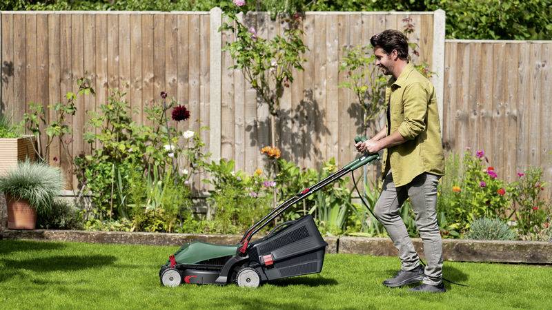 Ein Mann in einem Garten mäht den Rasen mit einem grünen Rasenmäher. Im Hintergrund stehen bunte Blumen und ein Holzzaun.