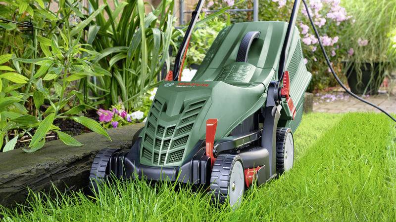 Ein grüner elektrischer Rasenmäher schneidet dichtes, grünes Gras in einem Garten mit blühenden Pflanzen im Hintergrund.