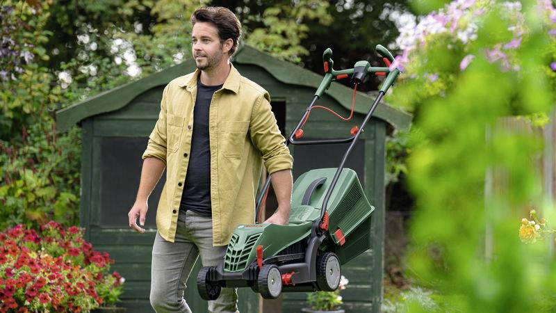 Ein Mann mäht mit einem grünen Rasenmäher den Rasen vor einem Gartenhaus, umgeben von blühenden Pflanzen.