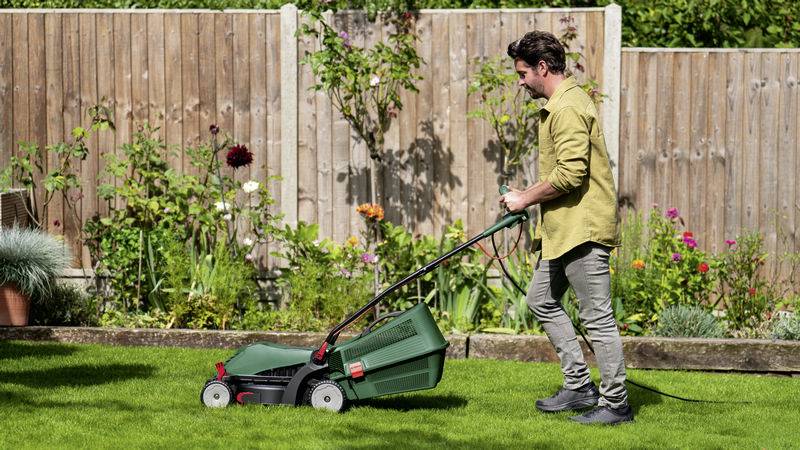 Ein Mann mäht mit einem Rasenmäher auf einer grünen Rasenfläche, umgeben von einem Holzzaun und bunten Blumen im Garten.