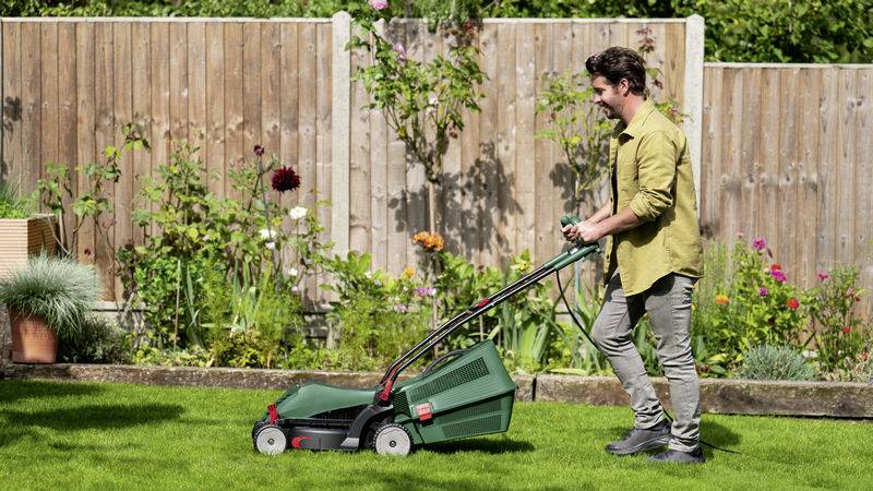 Ein Mann in einem Garten schiebt einen elektrischen Rasenmäher über einen grünen Rasen. Hintergrund mit Holzzaun und bunten Blumen.