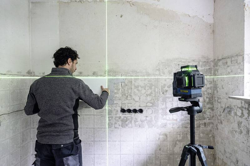 Ein Mann richtet Fliesen mit einem Kreuzlinienlaser an der Wand eines renovierungsbedürftigen Raums aus.