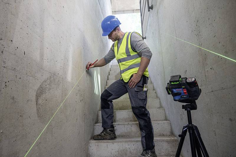 Ein Bauarbeiter mit blauer Schutzhelm und gelber Warnweste markiert mit einem Lasergerät präzise eine Wand in einem Treppenhaus.