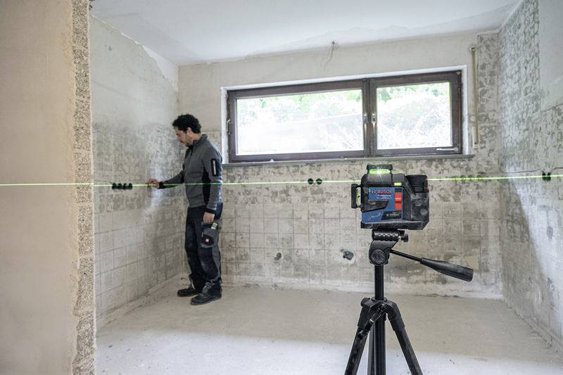 Ein Handwerker misst in einem leeren Raum mit gefliesten Wänden und Fenstern die Wand mit einem Laser. Ein Lasergerät steht auf einem Stativ im Vordergrund.