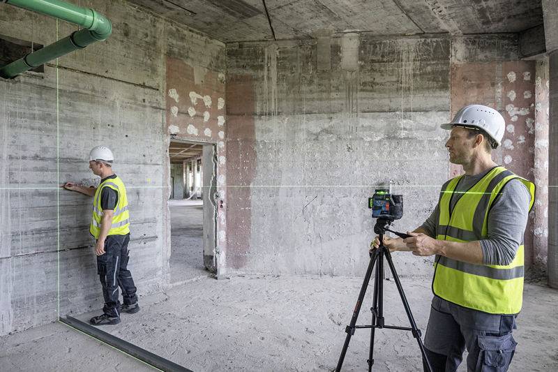Zwei Bauarbeiter in Sicherheitskleidung und Helmen arbeiten in einem unfertigen Gebäude. Einer benutzt ein Lasergerät zur Vermessung.