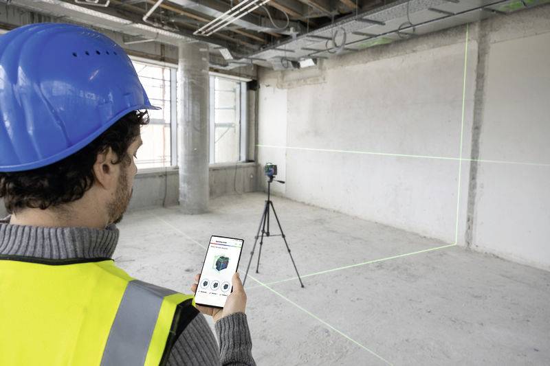 Ein Bauarbeiter mit Schutzhelm und Weste benutzt ein Smartphone, um ein Vermessungsgerät auf einem Stativ in einem leeren Raum zu steuern.