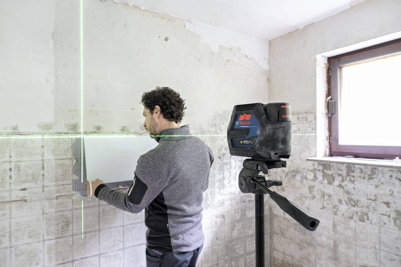 Ein Mann platziert Fliesen mit Hilfe eines an der Wand montierten Lasernivelliergeräts in einem unfertigen Raum.
