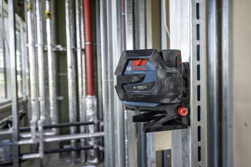 Ein Bosch Kreuzlinienlaser hängt an einem Metallgestell in einer Bauumgebung. Im Hintergrund sind Rohrleitungen sichtbar.
