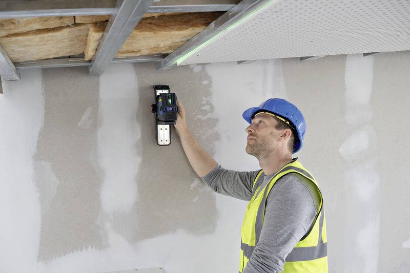 Ein Bauarbeiter in Warnweste und Schutzhelm benutzt ein digitales Messgerät an einer Wand, um Maße zu nehmen.