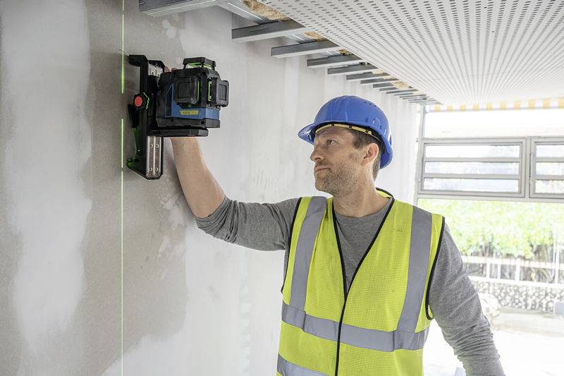 Ein Bauarbeiter trägt einen blauen Helm und eine gelbe Warnweste. Er arbeitet mit einem Laser-Werkzeug an einer weißen Wand.