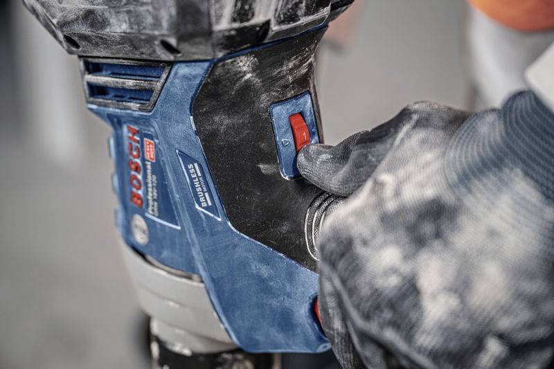 Hand mit Handschuh benutzt Elektrowerkzeug, blaue und schwarze Farben mit rotem Schalter sichtbar, verwendet in Werkstattumgebung.