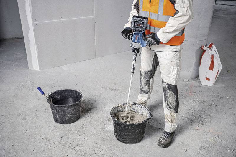 Eine Person in Arbeitskleidung mischt Zement in einem Eimer mit einem Elektrowerkzeug auf einer Baustelle. Ein weiterer Eimer steht daneben.