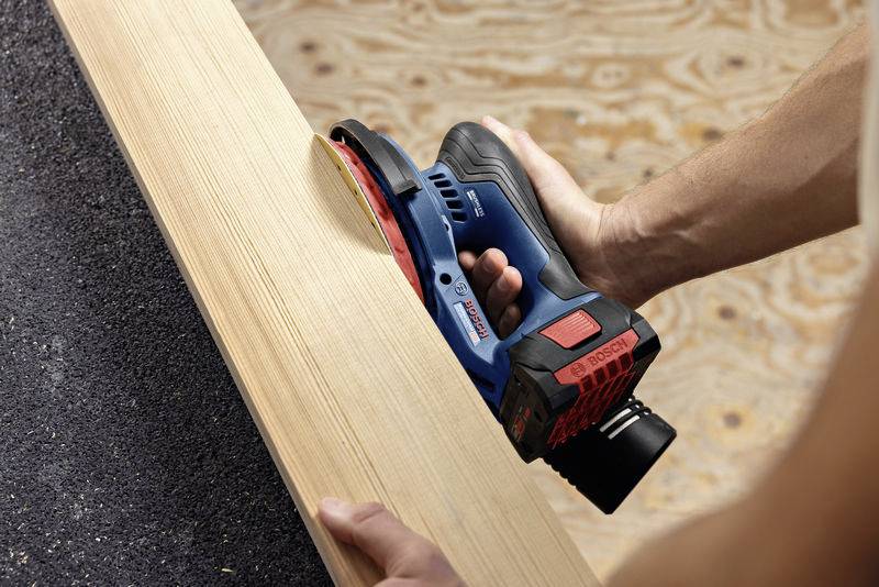 Eine Person schleift ein Holzbrett mit einem Elektrowerkzeug auf einer Werkbank. Das Brett ist teilweise geglättet.