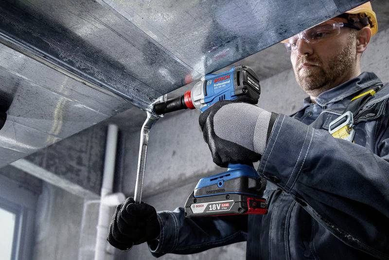 Ein Bauarbeiter in Schutzkleidung verwendet eine blaue Akku-Bohrmaschine, um Metall an der Decke zu befestigen, in einem industriellen Umfeld.