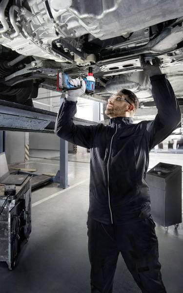 Mechaniker in Werkstatt repariert Auto von unten. Er trägt schwarze Arbeitskleidung und benutzt ein elektrisches Werkzeug.