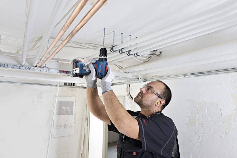 Ein Arbeiter bohrt mit einer elektrischen Bohrmaschine in die Decke, um Rohre zu montieren. Er trägt Schutzbrille und Handschuhe.