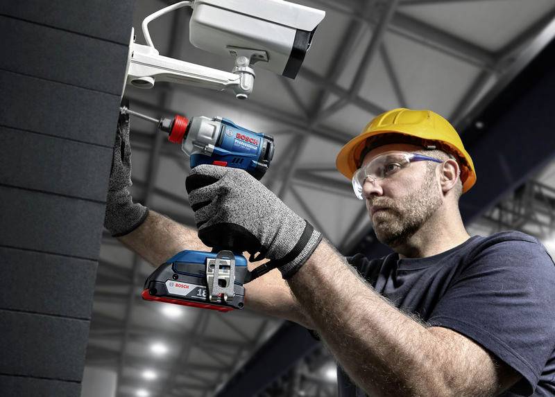 Ein Mann in Schutzkleidung installiert eine Überwachungskamera an einer Wand. Er benutzt einen blauen Akkuschrauber für die Montage.